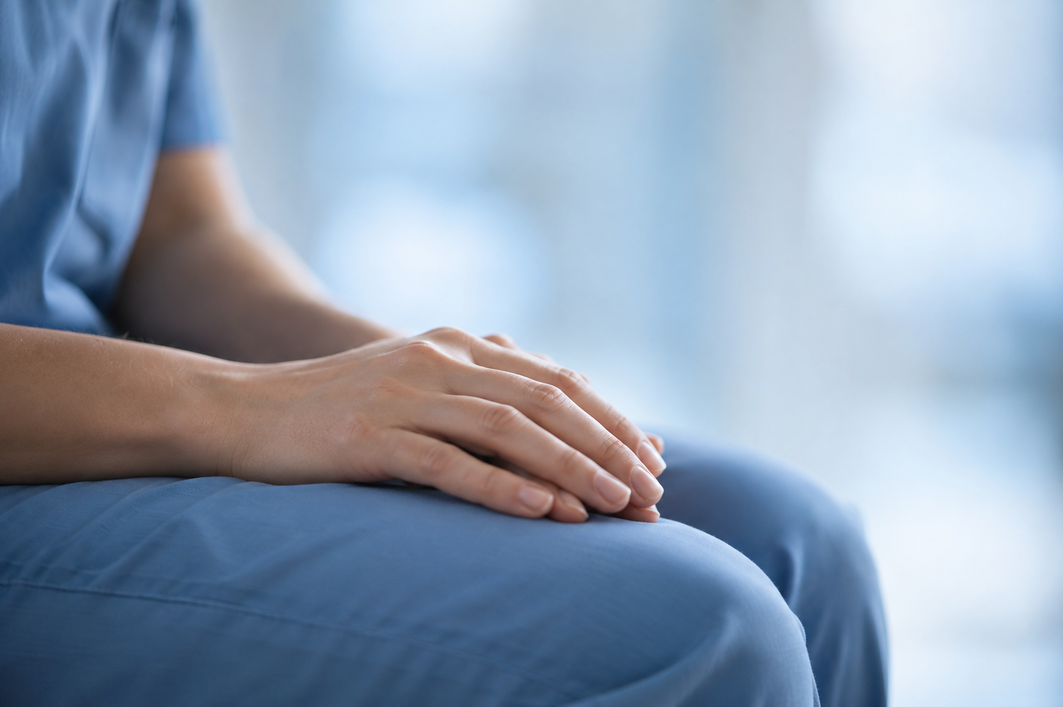 A person in blue scrubs sitting with their hands resting together on their knee, in soft light